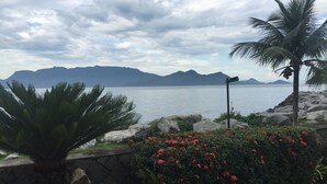 A coastal landscape with a view of mountains, a body of water, and a stone wall with red flowers. - Paradise House - Angra dos Reis (Conceição de Jacareí)