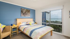 Une chambre avec une grande fenêtre, un lit recouvert d’une couverture aux couleurs vives, une chaise en osier et une table de chevet avec une lampe.
