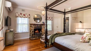 Una cama con dosel, una chimenea de piedra, un televisor y un espejo en un dormitorio luminoso con pisos de madera.