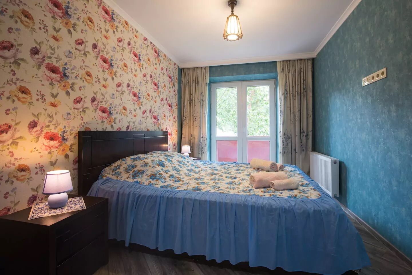 Une chambre à coucher avec un papier peint à motifs floraux, une couvre-lit bleue, une table de chevet avec une lampe et une fenêtre avec des rideaux.