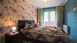 Een slaapkamer met bloemenbehang, een bed met een geborduurd sprei, een nachtkastje met een lamp en een raam met gordijnen.