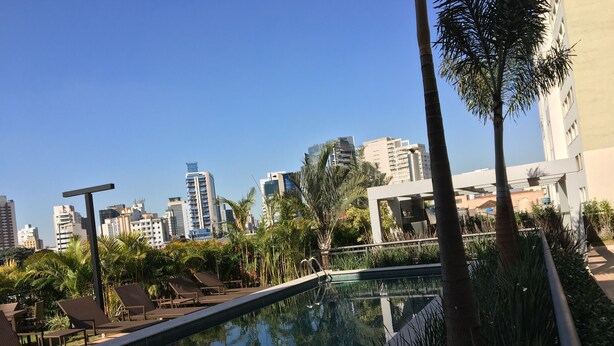 A rooftop pool with lounge chairs and palm trees, surrounded by modern buildings. - Brand new, cozy apartment, close to the subway, fast Wi-Fi, Netflix.  (Pinheiros)
