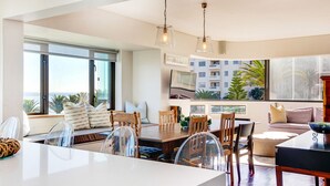 Una sala de estar moderna con comedor, ventanas amplias y vistas a un edificio y palmeras.