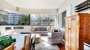 Una sala de estar moderna con techo curvo, un sofá gris, una mesa de comedor de madera y un gran ventanal con vistas a edificios.