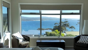 A bedroom with a large window offering a view of the sea and distant hills. There is a bed with white linens and two armchairs with patterned cushions. - FIVE BEACHES BEACH HOUSE Bay of Fires (Binalong Bay)