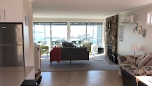 A modern living space with a kitchen, dining area, and seating by a large window overlooking a balcony. - FIVE BEACHES BEACH HOUSE Bay of Fires (Binalong Bay)