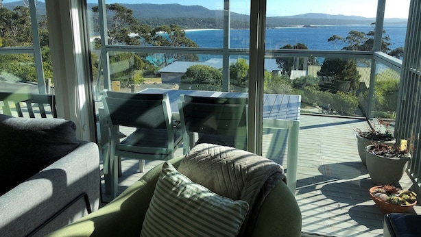 A sunlit deck with a view of the sea and hills, furnished with chairs and a table, and potted plants. - FIVE BEACHES BEACH HOUSE Bay of Fires (Binalong Bay)