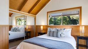 A bedroom with a large bed, a mirror, a window with a view of trees, and a wooden ceiling. - Windsong at Ravensbourne Escape 3 Bedrooms (Ravensbourne)