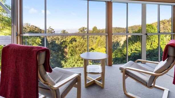 A room with two lounge chairs, a small table, and large windows offering a view of a forest and distant hills. - Windsong at Ravensbourne Escape 3 Bedrooms (Ravensbourne)
