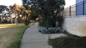 Stroll along the serene grassy path lined with trees and a metal fence, adjacent to a modern building with a concrete facade.
