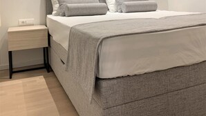 A modern bedroom with a grey bed, white pillows, a grey headboard, a bedside table with a lamp, and a wall-mounted abstract art piece. - Standard One-Bedroom Apartment With Balcony And Swimming Pool (Trogir)