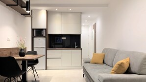 A modern kitchen with white cabinets, a built-in microwave, and a TV. There is a dining area with a table and chairs, and a gray sofa with yellow cushions. - Standard One-Bedroom Apartment With Balcony And Swimming Pool (Trogir)
