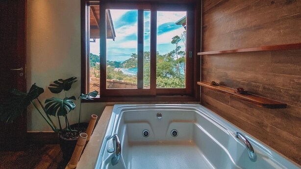Une salle de bain moderne avec une grande baignoire, un lambris en bois, une fenêtre donnant sur un paysage côtier et une étagère ornée d’objets décoratifs.