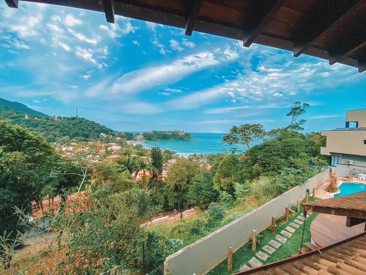 A view from a balcony overlooking a coastal area with a swimming pool, lush greenery, and a distant view of the sea and hills.