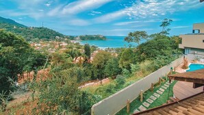 Une vue depuis un balcon donnant sur une zone côtière, avec une piscine, une végétation luxuriante et une vue lointaine sur la mer et les collines.