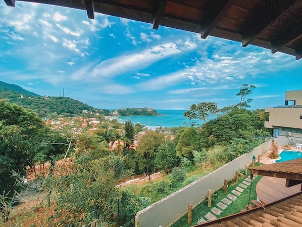 A view from a balcony overlooking a coastal area with a swimming pool, lush greenery, and a distant view of the sea and hills.