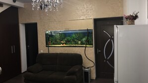 Un salon avec un canapé foncé, un lustre, un aquarium et un réfrigérateur.