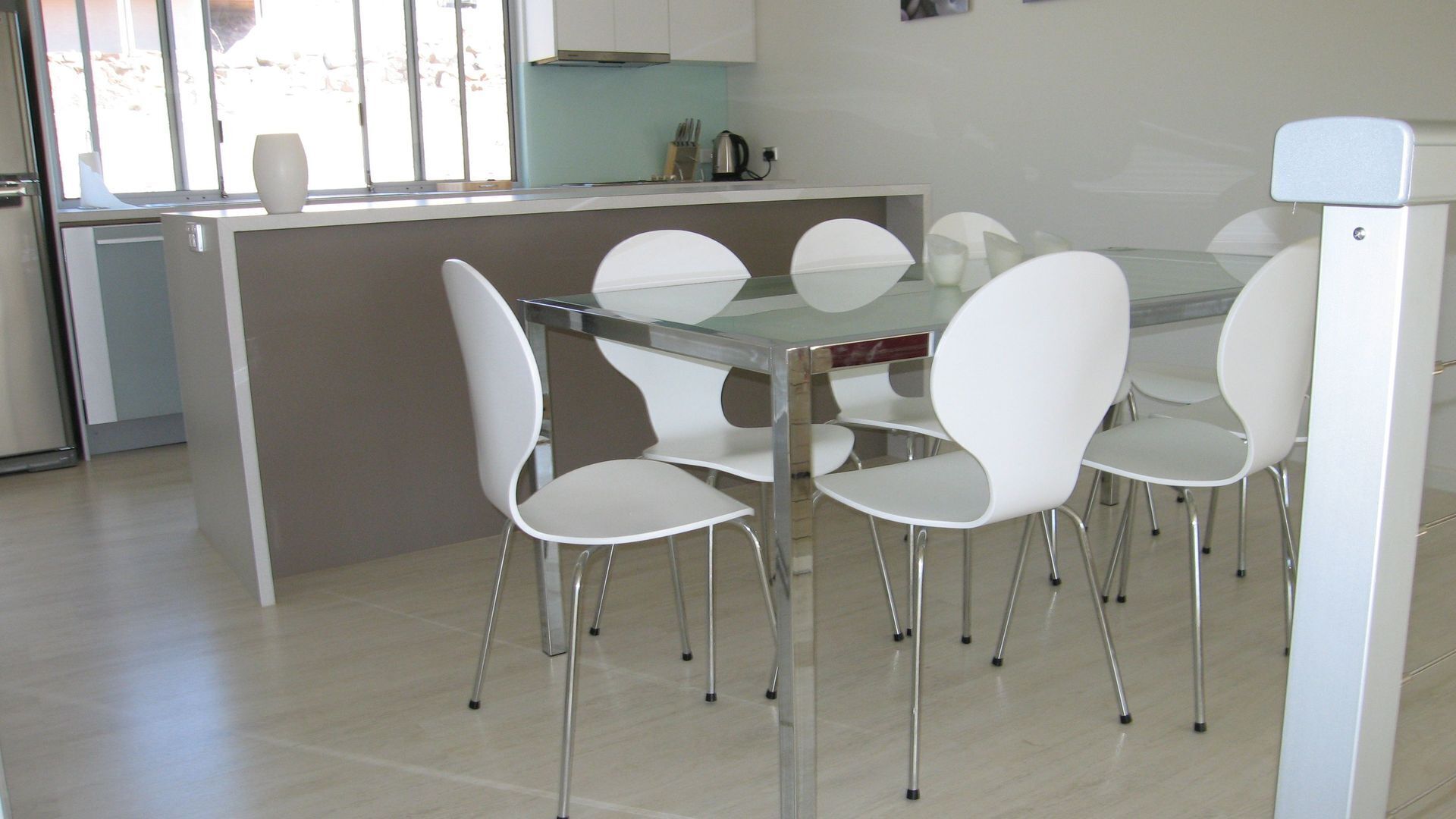 A modern kitchen with a glass dining table surrounded by white chairs.