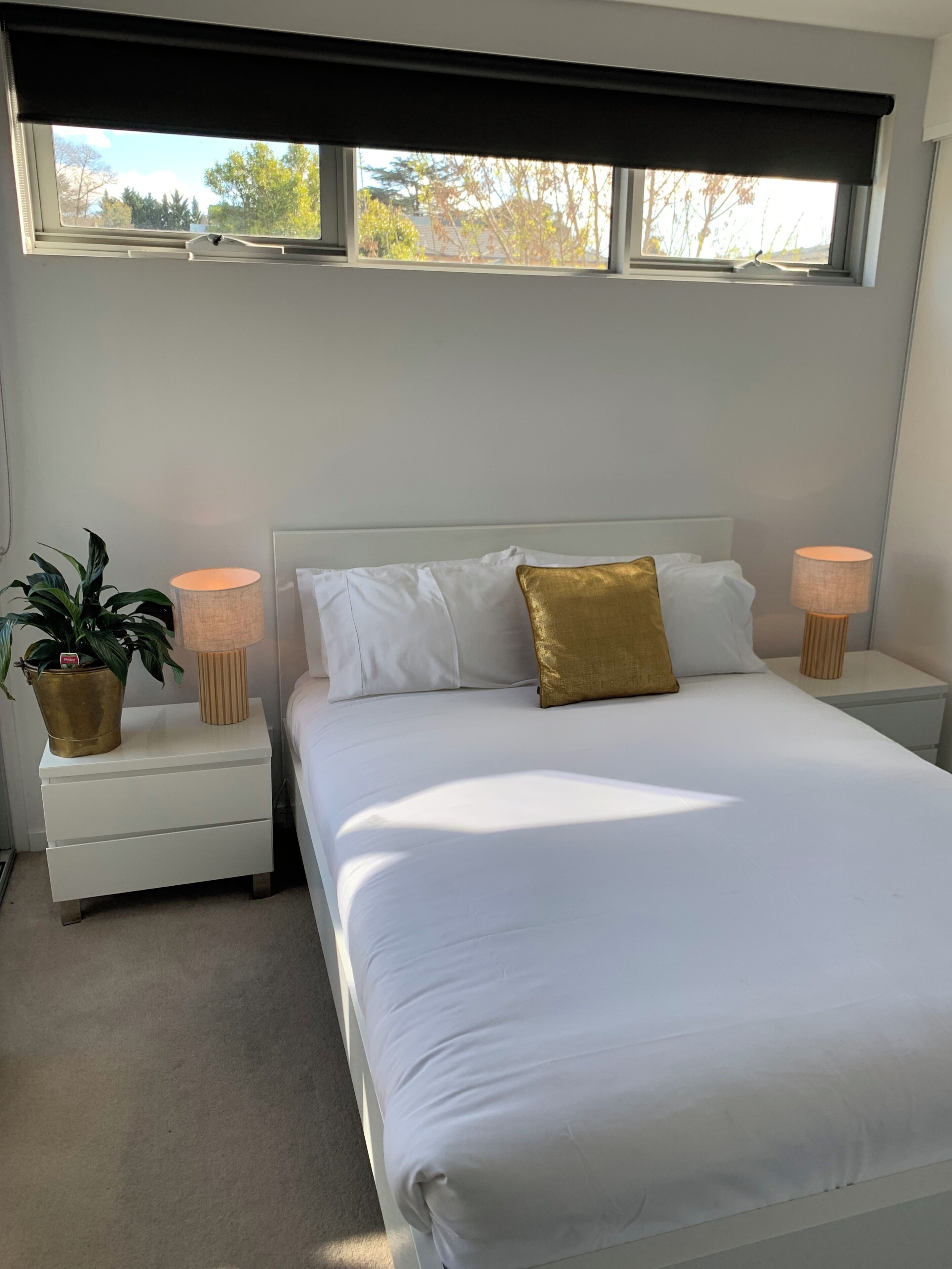 A bedroom with a large bed, two bedside tables, a window with a blind, and a potted plant.