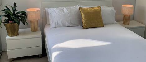 A bedroom with a large bed, two bedside tables, a window with a blind, and a potted plant.