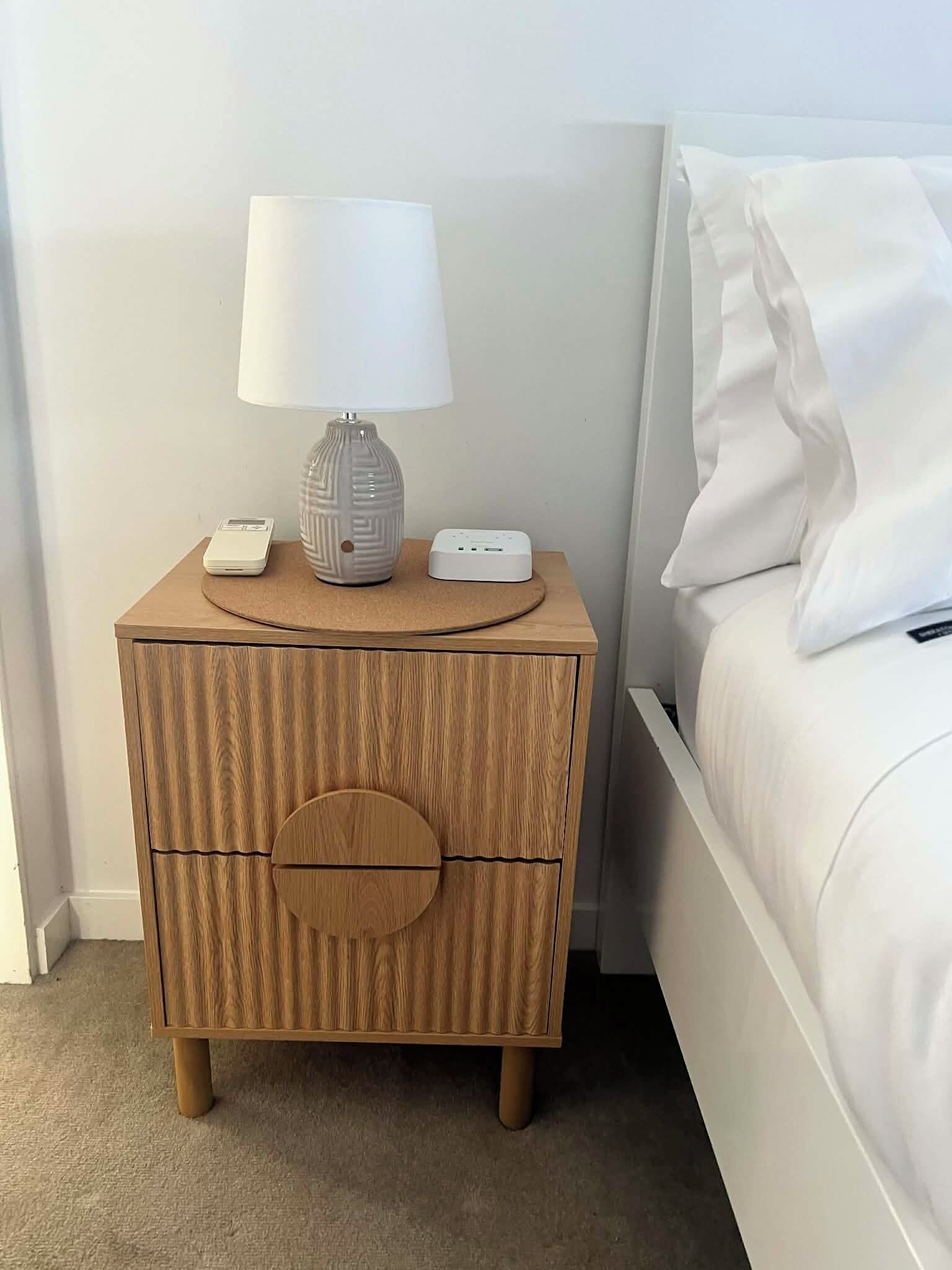 A wooden nightstand with a lamp, a power strip, and a small device.