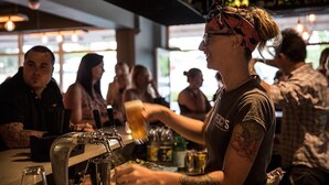 Un barman sert une boisson dans un bar, avec des clients assis à l'arrière-plan.