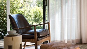 Een moderne kamer met een lederen fauteuil, een houten krukje en uitzicht op groen door een raam.