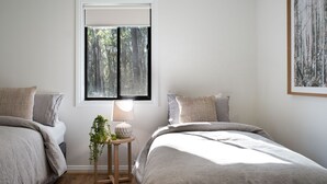Een slaapkamer met twee bedden, een raam met uitzicht op bomen, een nachtkastje met een lamp en een lijst met een afbeelding aan de muur.