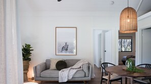 Uma sala de estar com sofá cinzento, um quadro emoldurado de cangurus, uma luminária pendente de fibra e um espaço de jantar com mesa e cadeiras de madeira.