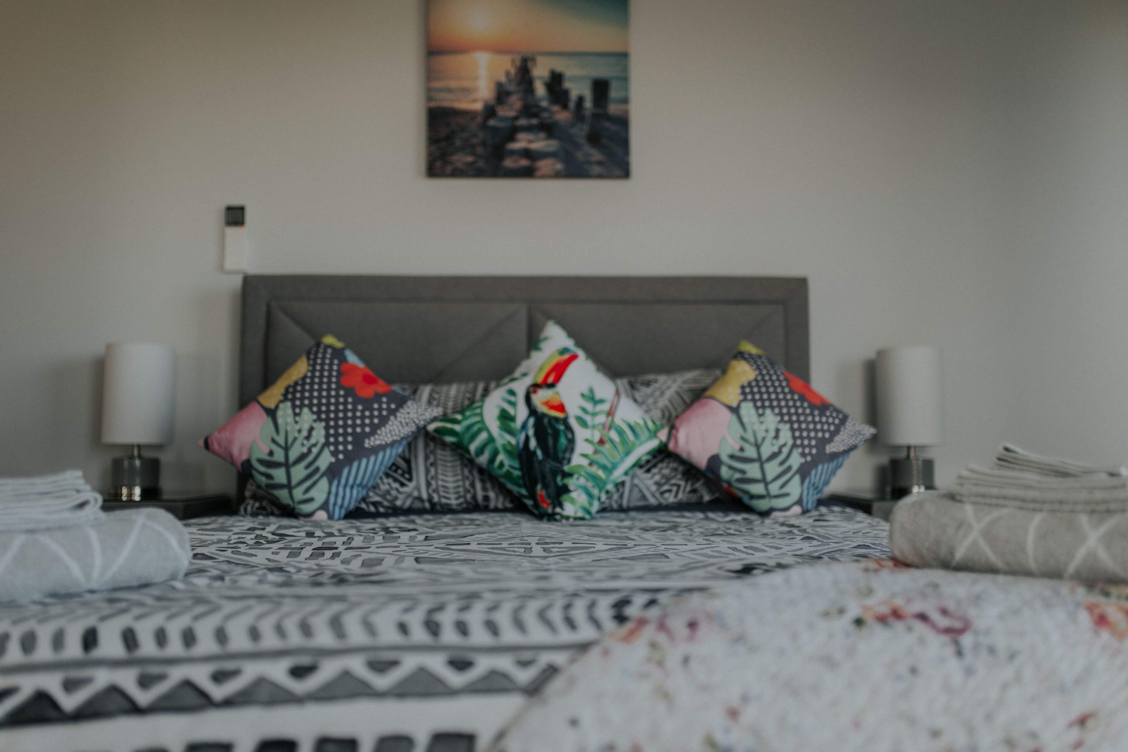 A neatly made bed with patterned bedding, a cityscape painting on the wall, and a nightstand with a lamp.