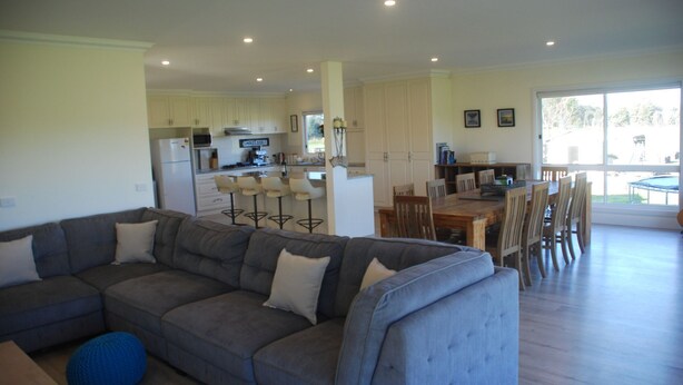 A modern living room with a grey sofa, a dining area with wooden chairs, and a kitchen in the background. - Jackaleah Farm  Stay Oxley (Oxley)
