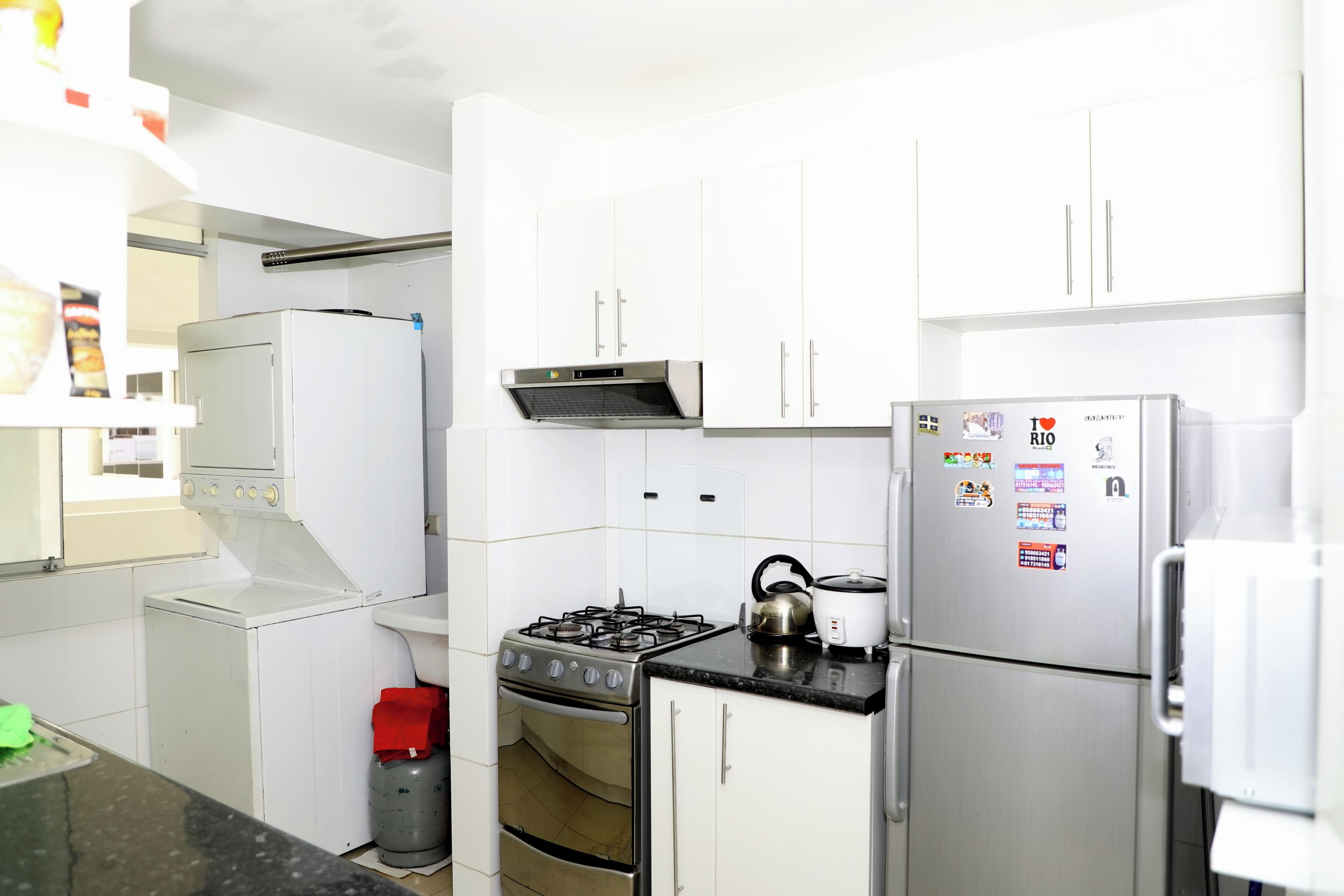 Une cuisine avec des meubles blancs, une cuisinière à gaz, un four encastré, un réfrigérateur et une bouteille de gaz.