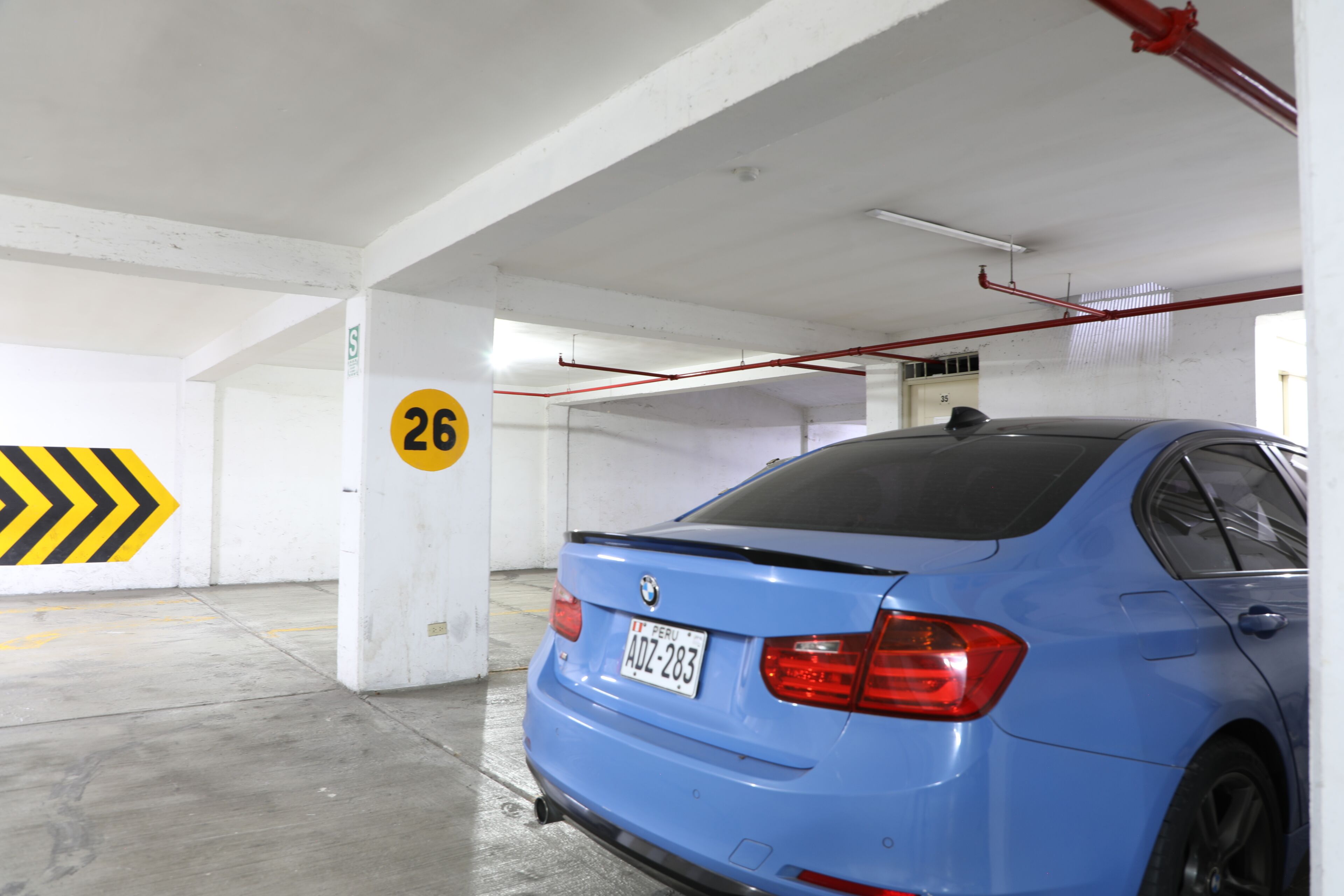 Un BMW azul estacionado en un garaje con un letrero direccional amarillo y negro, pilares de concreto y una tubería roja en el techo.