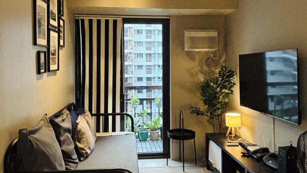Un espace de vie compact comprenant un lit, une télévision, un bureau avec une lampe et un balcon agrémenté de plantes.
