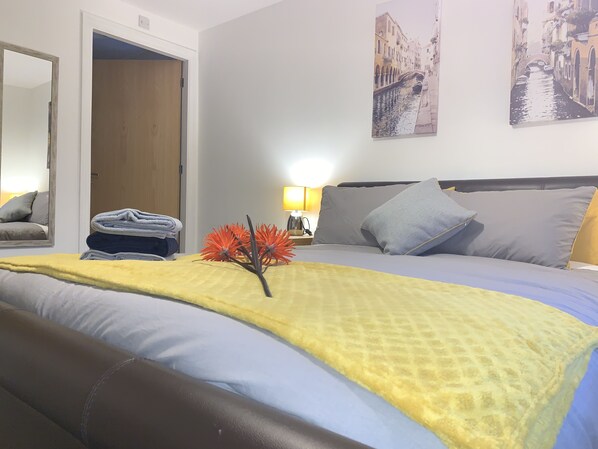 A hotel room with a bed, a bedside table with a lamp, a mirror, and two paintings on the wall.