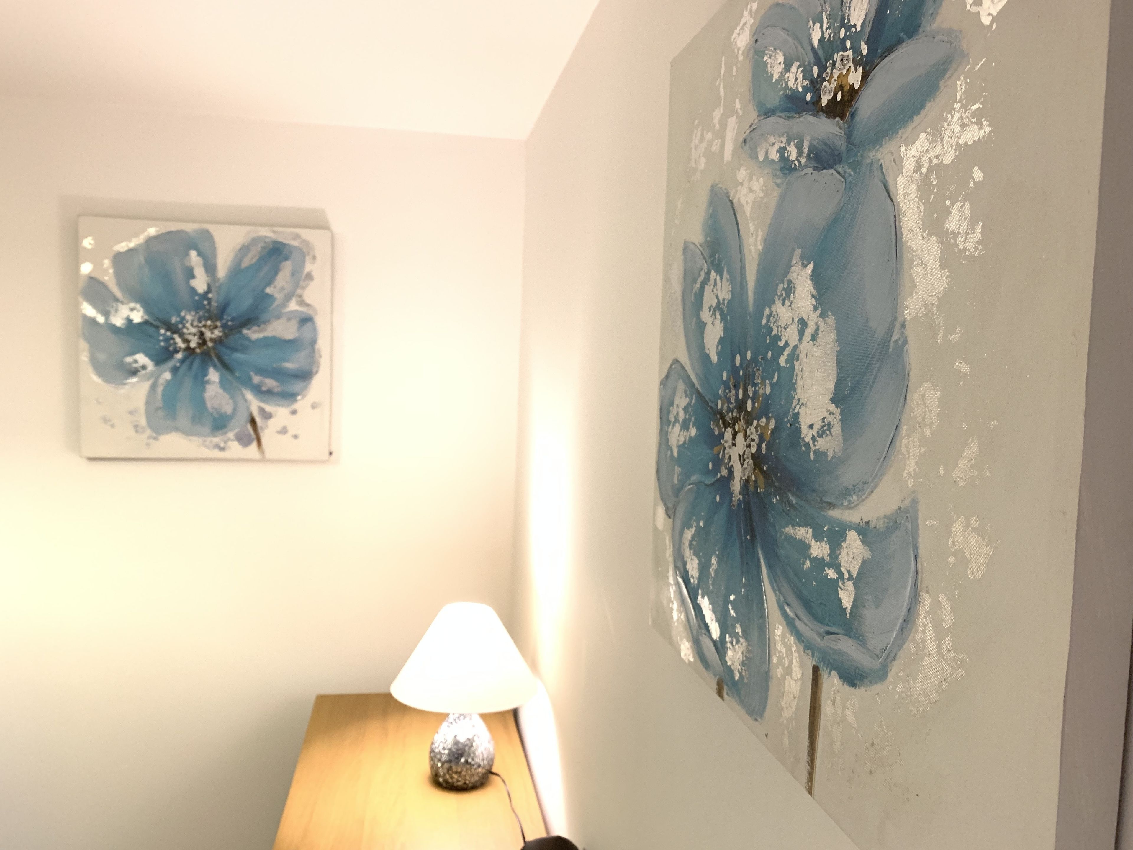 A room with a large floral painting on the wall, a small table with a lamp, and a framed picture above the table.