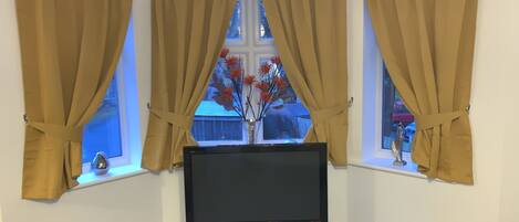 A living room with a flat-screen TV on a black stand, beige curtains, and a window with a view of a house and a tree.
