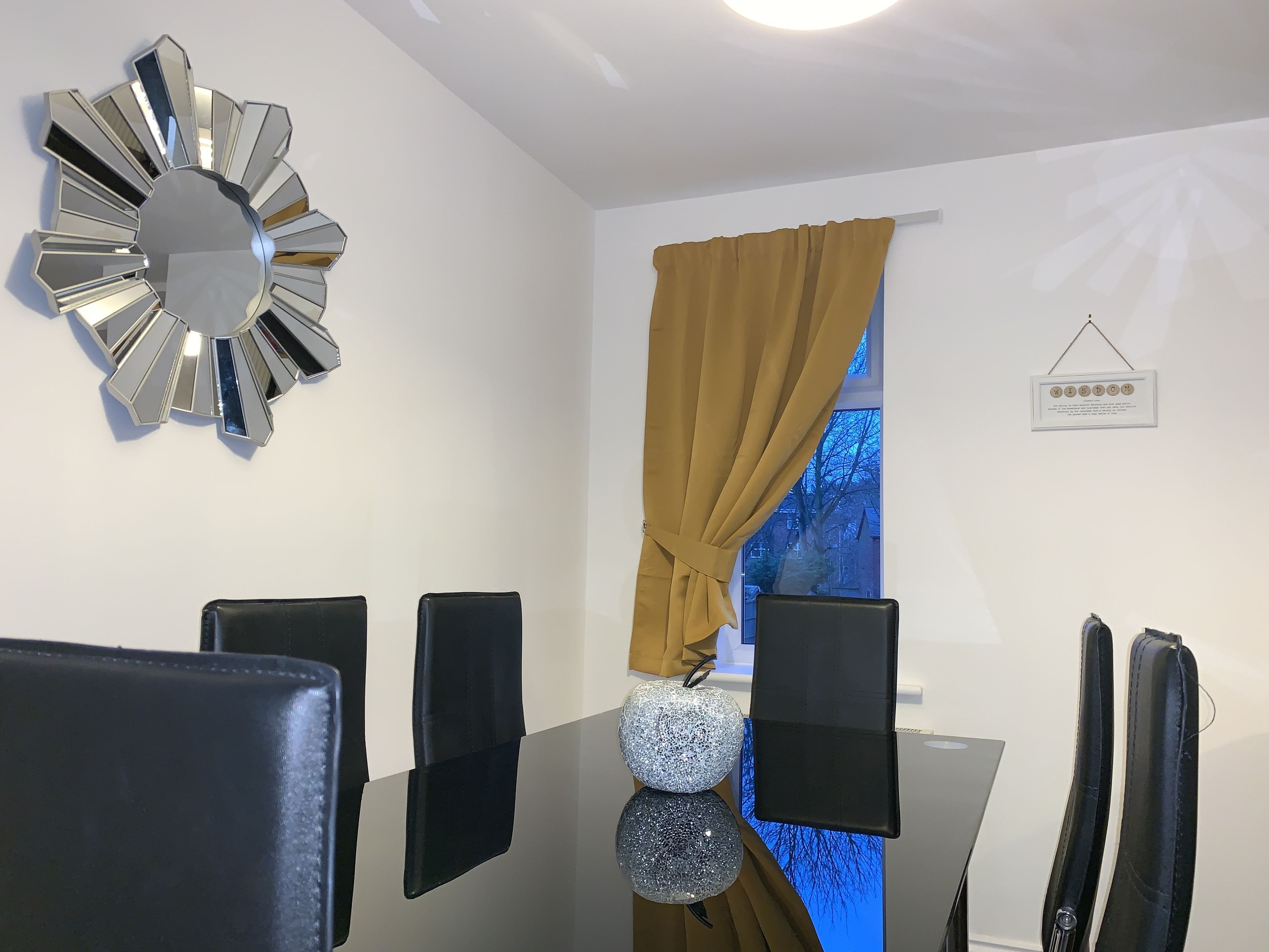 A modern dining area with a unique metallic wall clock, a table with black chairs, and a window with a view of trees.