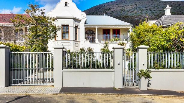 Uma casa de estilo colonial branca com varanda, cercada por um muro branco e uma montanha ao fundo.