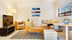 Uma sala de estar moderna com sofá, mesa de centro e aparador para TV. Há três quadros na parede e uma janela com persianas.
