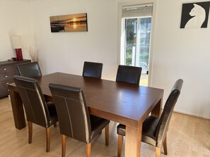 A dining area with a wooden table and six chairs, a painting on the wall, and a window with blinds. - Relax at "Kangaroo Pause" @ Clayton Bay (Clayton Bay)
