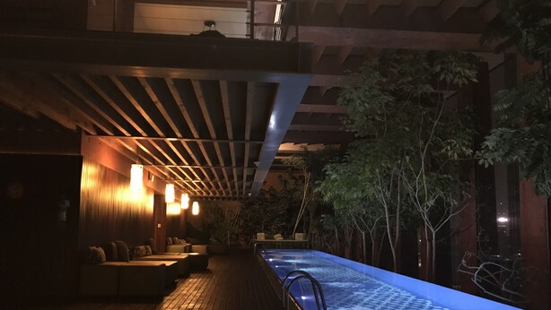 Een zwembadruimte met weinig licht, voorzien van een houten terras, loungezitplaatsen en een decoratieve poolrand.