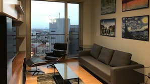 Een moderne woonkamer met een bank, een stoel, een glazen salontafel en een groot raam met uitzicht op de stad.