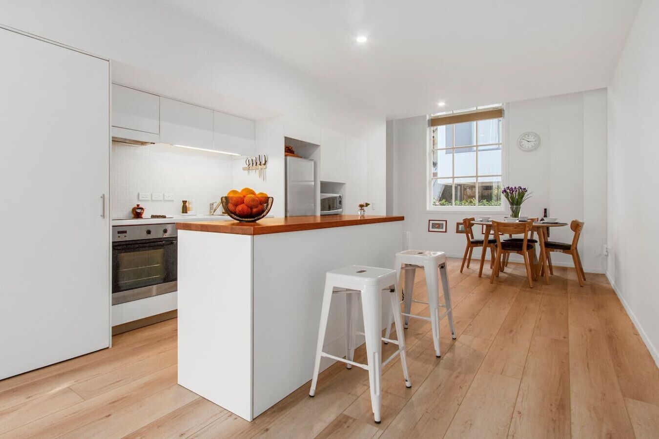 A modern kitchen with white cabinetry, a wooden countertop, and a built-in oven. There is a dining area with wooden chairs and a table, and a window providing natural light.