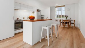 A modern kitchen with white cabinetry, a wooden countertop, and a built-in oven. There is a dining area with wooden chairs and a table, and a window providing natural light. - City Fringe Converted Industrial Townhouse (Auckland)