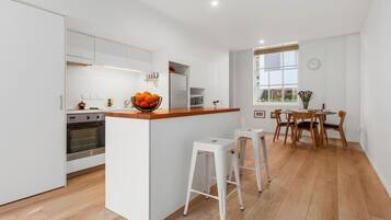 A modern kitchen with white cabinetry, a wooden countertop, and a built-in oven. There is a dining area with wooden chairs and a table, and a window providing natural light.