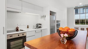 A modern kitchen with white cabinetry, stainless steel appliances, and a wooden dining table with a fruit bowl. - City Fringe Converted Industrial Townhouse (Auckland)