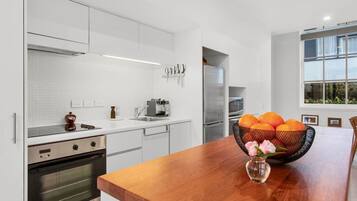 A modern kitchen with white cabinetry, stainless steel appliances, and a wooden dining table with a fruit bowl.
