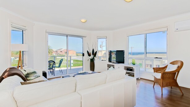 A spacious living room with a white sofa, a television, and a large window offering a view of the sea. - Sunset at Snapper Point ~ Aldinga Beach - Modern Coastal Beauty with Large Balcony and Sea Views (Aldinga Beach)