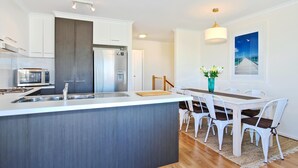 A modern kitchen with white cabinets, a stainless steel refrigerator, a microwave, and a sink. There is a dining area with a white table and chairs, a vase with flowers, and a painting of a pier on the wall. - Sunset at Snapper Point ~ Aldinga Beach - Modern Coastal Beauty with Large Balcony and Sea Views (Aldinga Beach)